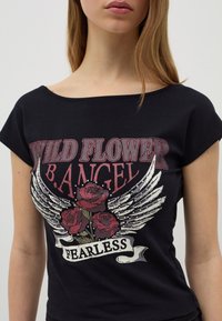 Camiseta negra de manga corta con un diseño gráfico que incluye "WILD FLOWER B.ANGEL", alas y rosas rojas, con una pancarta de "FEARLESS" debajo.