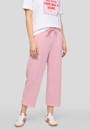 Frau trägt weißes T-Shirt mit rotem Text, rosa-weiß gestreifte, weit geschnittene Hose und weiße Sneaker mit roten Streifen.