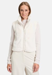 Biała kamizelka z futerkiem syntetycznym, z wysokim kołnierzem, zapięciem na zamek błyskawiczny i luznym krojem. Nosi się ją na jasnym swetrze, zestawiona z kremowymi spodniami.