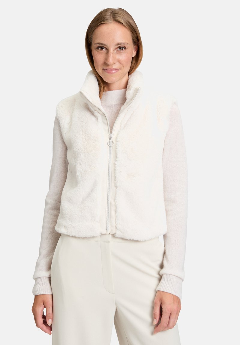 Biała kamizelka z futerkiem syntetycznym, z wysokim kołnierzem, zapięciem na zamek błyskawiczny i luznym krojem. Nosi się ją na jasnym swetrze, zestawiona z kremowymi spodniami.