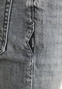 Jeans de mezclilla gris con una textura sutil, con una etiqueta negra que dice "CHASIN'" visible en la esquina del bolsillo.