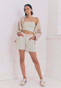 Top a tubo verde chiaro, camicia oversize a righe beige e bianche, pantaloncini verdi chiaro con tasche, sneaker beige con dettagli blu navy.