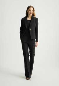 Completo tailored nero con giacca a un bottone e lapels affilati, pantaloni dritti e una maglia nera aderente sotto.