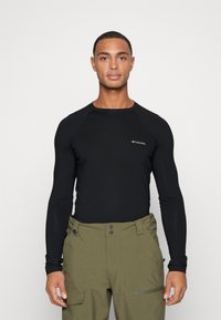 Camisa negra de manga larga con mangas raglán, que presenta un pequeño logo de Columbia en el pecho, combinada con pantalones verde oliva.