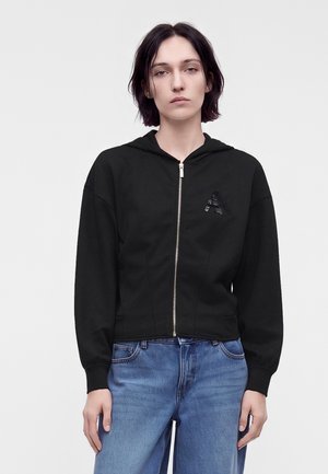 Ung person med kort mørkt hår iført en sort hættetrøje med lynlås og hætte samt blå jeans, der står foran en ensfarvet baggrund.