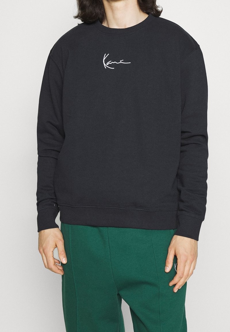 Sudadera negra hecha de material suave con mangas largas, puños acanalados y cuello redondo. Presenta un logo blanco bordado en el pecho.