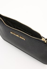 Czarna skórzana kopertówka o teksturowanym wykończeniu, złoty zamek błyskawiczny i wytłoczone logo "MICHAEL KORS" na przodzie. Odsłonięty zamek ukazuje wnętrze.