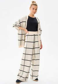 Beige gebreide cardigan met zwart ruitpatroon, in combinatie met bijpassende wijde broek. Zwart topje eronder. Losse pasvorm, grote knopen op de cardigan.