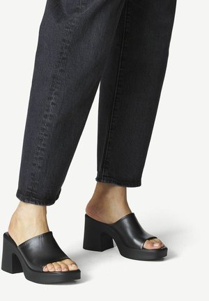 Mules noires à talons blocs ouverts, avec une tige en cuir lisse et une semelle en caoutchouc épaisse, assortis à un pantalon en denim sombre à jambe fuselée.