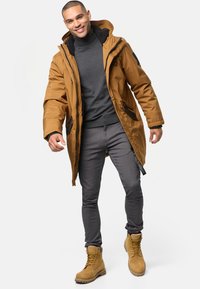 Chaqueta de plumas khaki con acentos negros, suéter gris de cuello alto, pantalones grises de corte ajustado y botas color trigo con cordones y suelas robustas.