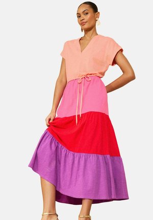 Žena v vrstvených ružových, červených a fialových midi šatách s krátkymi rukávmi a viazaním v páse, pózuje jednou rukou na bok a drží lem sukne.