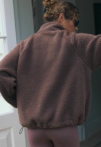 Pullover in pile marrone con colletto alto, vita elastica e superficie texturizzata. Presenta una vestibilità rilassata e cuciture delle spalle arrotondate.