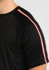Camiseta atlética de manga corta en tejido negro con franjas laterales rosas. El diseño cuenta con un panel de malla transpirable en el hombro.