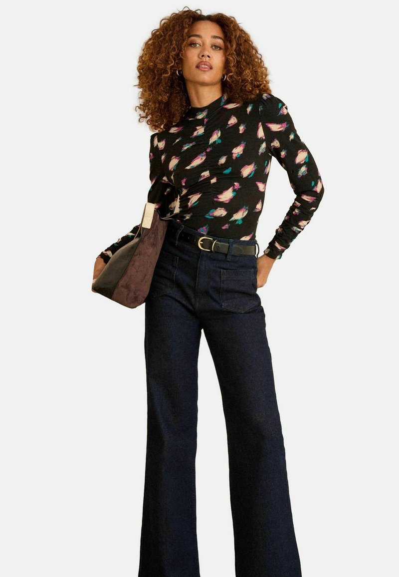 Hosszú ujjú fekete blúz színes tollmintával, sötét farmer harangnadrággal párosítva és egy barna velúrbőr táskával, aranyszínű övvel.