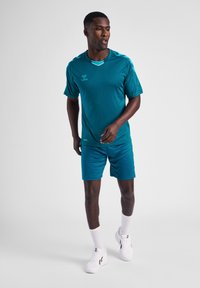 Turquoise atletisk skjorte og shorts i blødt, fugttransportende stof, med V-hals, logo-detalje og kontrastkant, parret med hvide sneakers.