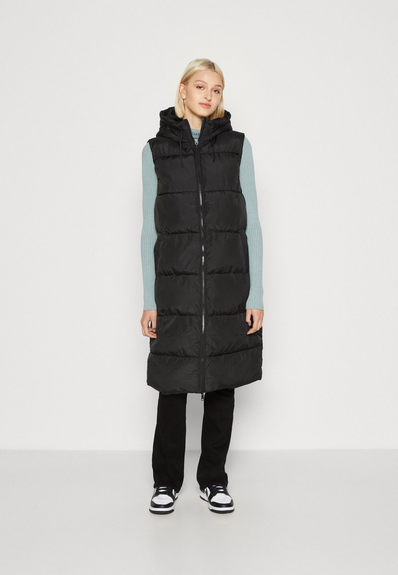 Zwarte gewatteerde vest met capuchon, met een rits aan de voorkant, een geregen design en zijzakken, gedragen over een lichtblauw sweater en zwarte broek.