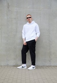 Sweatshirt à capuche blanche, pantalon cargo noir et baskets Nike blanches avec des accents noirs. Le modèle se tient contre un mur en béton gris.