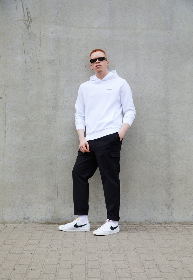 Sweatshirt à capuche blanche, pantalon cargo noir et baskets Nike blanches avec des accents noirs. Le modèle se tient contre un mur en béton gris.