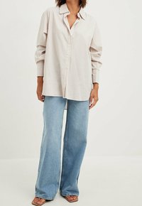 Light beige button-up shirt with a relaxed fit and long sleeves, paired with light blue flared jeans and flat sandals, against a neutral background.