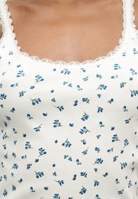 Κοντινό πλάνο κορμού που φοράει λευκό ριμπ αμάνικο τοπ με δαντέλα και μικρό μπλε floral σχέδιο.