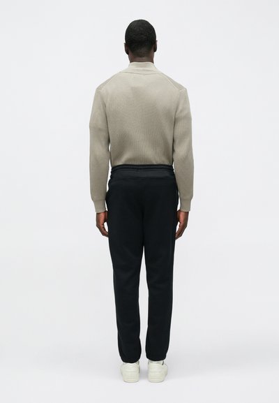 Μπεζ πλεκτό πουλόβερ με απλό γιακά, σε συνδυασμό με μαύρο φόρμα και λευκά αθλητικά παπούτσια, από οπίσθια όψη σε απλό φόντο.