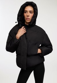 Czarna kurtka puchowa z kapturem, posiadająca pikowaną teksturę, przednie kieszenie oraz zamek błyskawiczny. Materiał wydaje się gładki i izolowany.