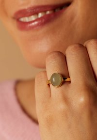 Goldring mit einem ovalen grünen Stein, umrandet von kleinen Perlen. Die Hand zeigt eine natürliche Hauttextur und subtile Nageldetails.