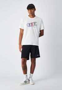 Camiseta de algodón blanca con gráfico del logo de Champion en varios colores, combinada con pantalones cortos negros. El modelo lleva zapatillas blancas y calcetines de estampado.