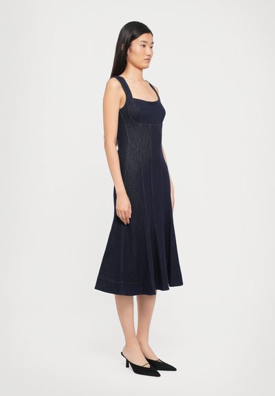 Marineblauwe denim jurk met een aangesloten lijfje en een uitlopende rok. Beschikt over brede schouderbanden en contrasterende stiksels. Zwarte puntige hakken maken de look compleet.