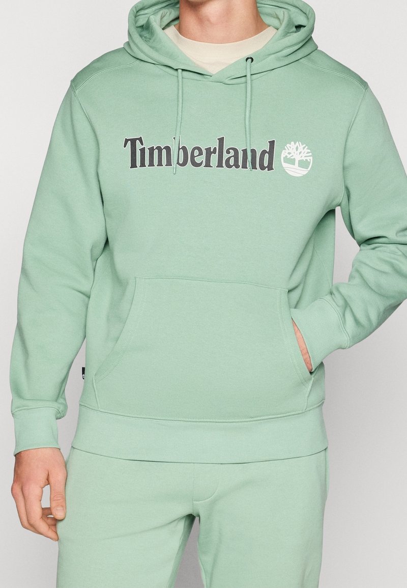 Homme portant un sweat à capuche Timberland vert clair avec poche avant et pantalon assorti, main dans la poche, sur fond uni.