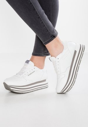 Osoba ubrana w czarne dżinsy i białe sneakersy na platformie z szarymi paskowanymi podeszwami, stojąca na gładkiej białej powierzchni.