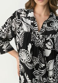 Túnica floral en blanco y negro con cuello camisero, mangas cortas y cintura fruncida, que presenta patrones audaces y una textura de tela suave.