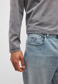 Ανοιχτό γκρι μπλούζα με μακρύ μανίκι φορεμένη με ξεθωρ ιασμένα μπλε τζιν που διαθέτουν κεντημένο το '73' στην πίσω τσέπη. Απαλή υφή και άνετη εφαρμογή.