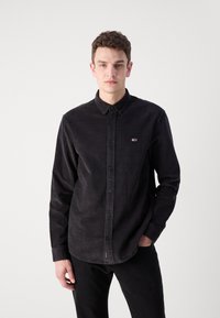 Camisa de pana negra con diseño de botones, cuello y parche con logo en el pecho. Las mangas largas y el bajo recto completan el look.