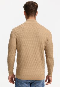 Pull en maille beige avec un motif en vagues texturé, col, poignets et ourlet côtelés, porté avec un jean en denim bleu.
