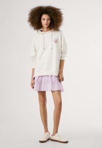 Lichtbeige hoodie met een trekkoord en geborduurd logo, gelaagd over een lavandelkleurige ruchesrok. Draag het met witte sneakers met een bruine zool.