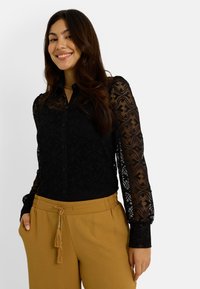 Blusa de encaje negra con mangas largas estampadas, frente abotonado, combinada con pantalones beiges de cintura elástica con detalle de borlas.