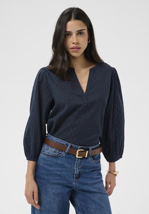 Young woman with long dark hair wearing a navy textured blouse, blue jeans, brown belt, and gold hoop earrings and bracelet against a white background.