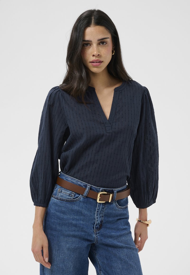Jeune femme aux longs cheveux foncés portant un chemisier texturé bleu marine, un jean bleu, une ceinture marron ainsi que des boucles d’oreilles et un bracelet en or sur fond blanc.