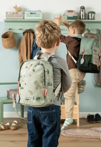 Deux jeunes enfants avec des sacs à dos accrochent des objets dans une entrée de maison avec des manteaux, des chaussures et des paniers de rangement.