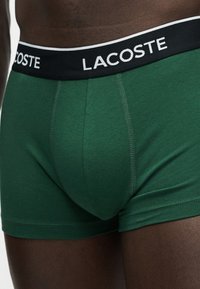 Gros plan d'une personne à la peau foncée portant un boxer vert avec une ceinture noire arborant le logo blanc Lacoste.