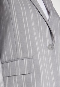 Szara marynarka w paski z klapami z wycięciem, zapinana na dwa guziki, z kieszenią na piersi. Tkanina ma subtelną fakturę i pionowe linie.