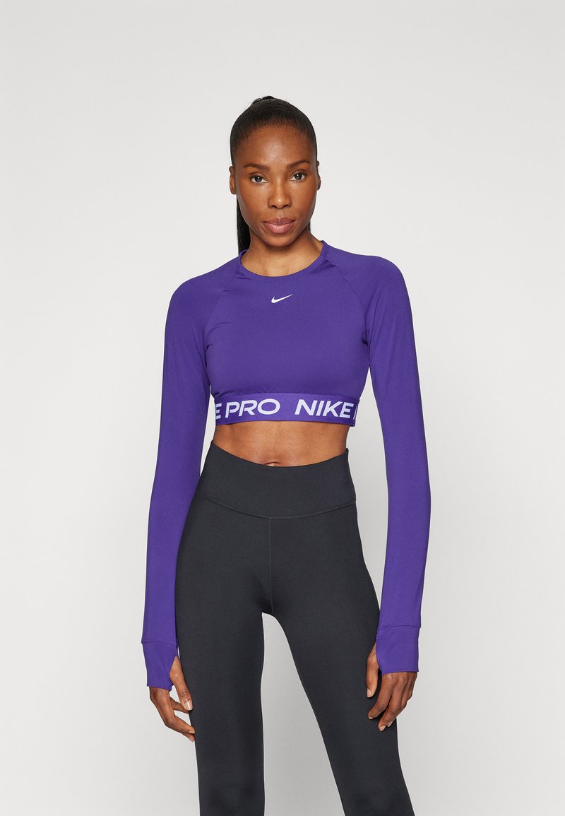 Lilla langermet crop top med rund hals, med en hvit merkebånd i livet og tettsittende svarte leggings. Glatt stofftekstur.