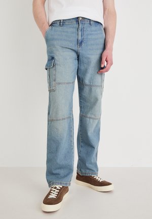 Light blue denim cargo pants with side pockets, a relaxed fit, and visible seam detailing. Paired with brown suede sneakers.