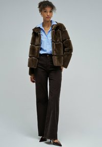 Chaqueta de piel sintética marrón con un diseño cuadrado, camisa azul de botones, pantalones de pana oscuros de pierna acampanada y tacones burdeos puntiagudos.