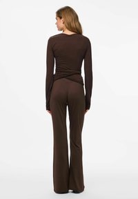 Sieviete ar muguru pret skatītāju, ģērbusies ietilpīgā tumši brūnā garajās piedurknēs topā ar savilktām detaļām un saskanīgās plati garās biksēs uz baltā fona.