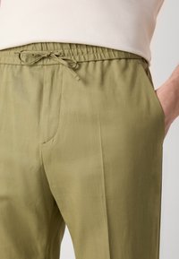 Olivgröna byxor med elastisk midja, justerbar snörning och sidofickor. Materialet verkar lätt och har en slät ytstruktur.