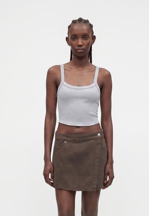 Femme aux cheveux tressés portant un débardeur court gris clair et une mini-jupe marron foncé, debout devant un fond uni.