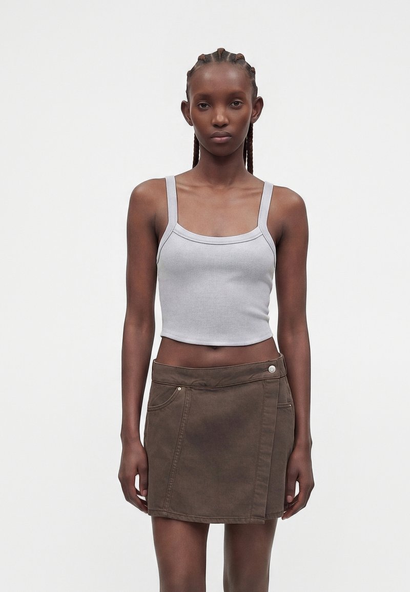 Femme aux cheveux tressés portant un débardeur court gris clair et une mini-jupe marron foncé, debout devant un fond uni.
