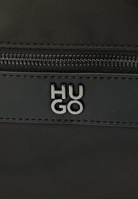 Μαύρη τσάντα από νάιλον με έντονο ασημί λογότυπο "HUGO", συνοδευόμενη από φερμουάρ και κομψή ραφή.